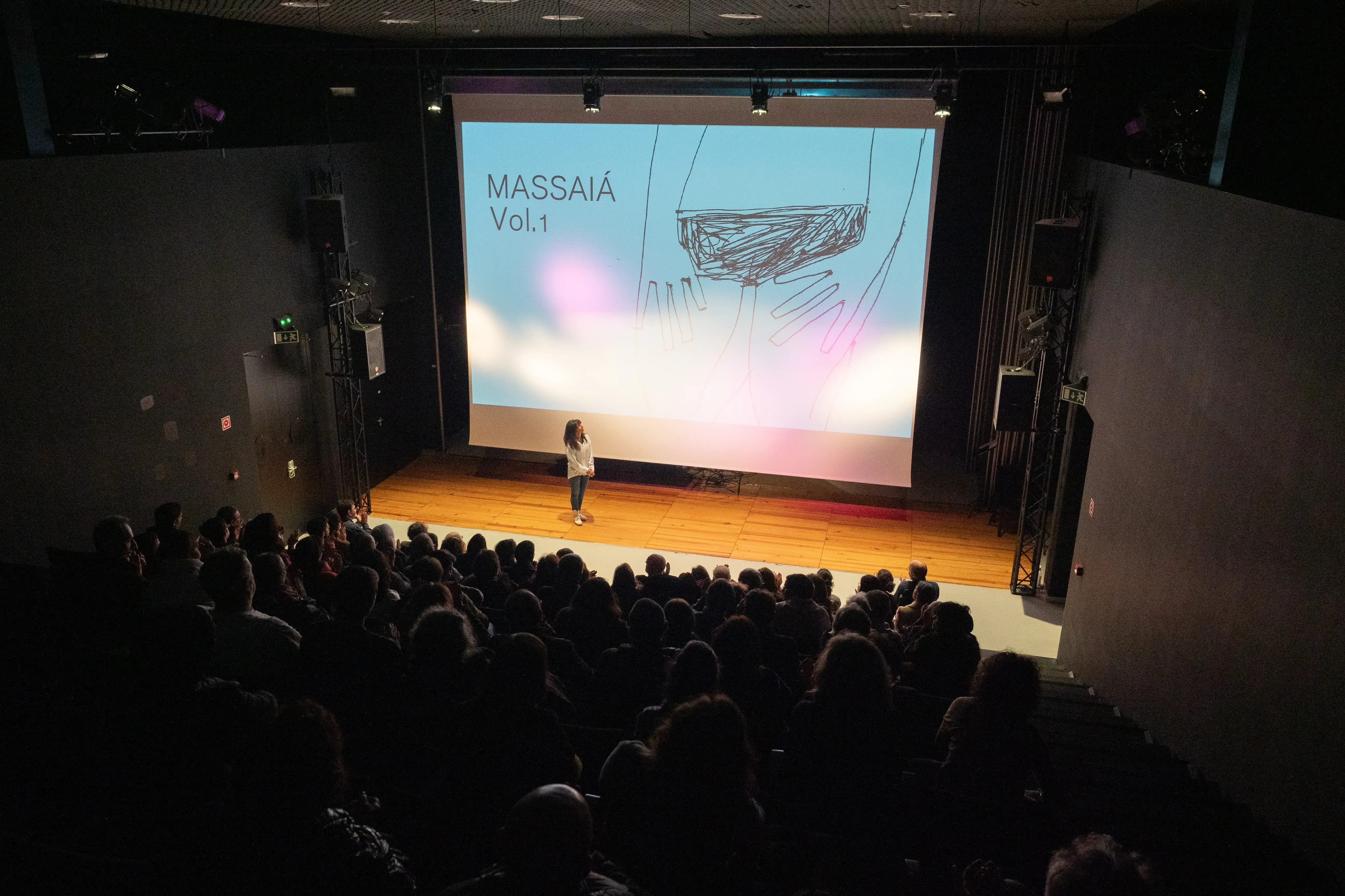 Ao fundo de uma plateia cheia, no palco, uma mulher em pé olhar para a projeção de uma ilustração a qual tem escrito ao lado “Massaiá Vol. 1”.